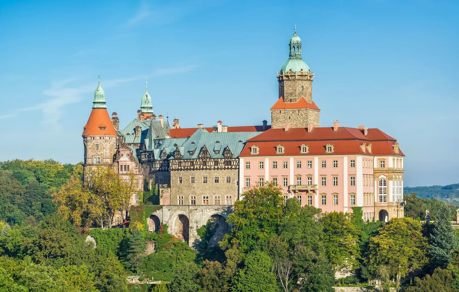 An ornate historic castle with a red roof and light pink facade, featuring several towers with green domed caps, stands majestically amidst green trees and foliage under a clear blue sky.